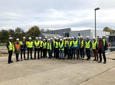 Eine Gruppe von etwa 20 Personen in Sicherheitswesten und weißen Schutzhelmen steht auf einem Industriegelände vor einem grauen Gebäude. Im Hintergrund sind Bäume und Baustellenmaterial zu sehen.