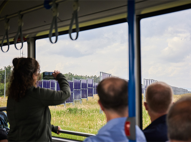 Das Innere eines Busses. Eine Frau hält ein Handy hoch und fotografiert durch das Fenster des Busses. Draußen sieht man eine Wiese mit Photovoltaikanlagen.