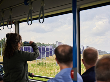 Das Innere eines Busses. Eine Frau hält ein Handy hoch und fotografiert durch das Fenster des Busses. Draußen sieht man eine Wiese mit Photovoltaikanlagen.