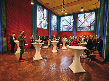 Menschen stehen an Stehtischen in einem großen Raum mit abstrakten Mustern auf dem Fußboden und an der Decke. Manche sitzen auf Hockern. Sie blicken zu einem Mann mit Mikrofon, der links steht und einen Anzug trägt.