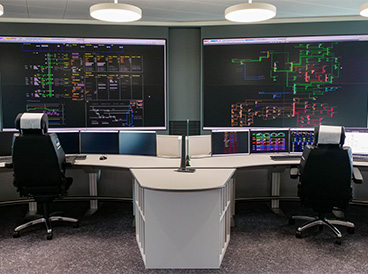Zwei große Kontrollmonitore zeigen farbige Diagramme und Tabellen. Davor stehen zwei Bürostühle und weitere kleinere Bildschirme.
