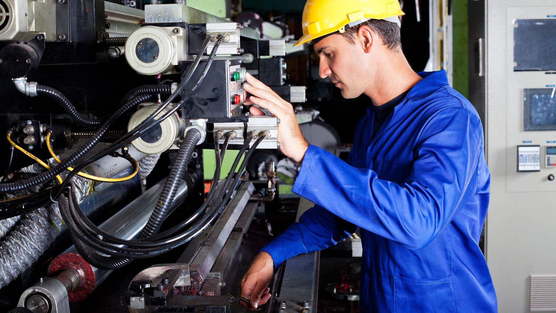 Ein Mann mit einem blauen, langärmligen Pullover und einem gelben Schutzhelm steht rechts von einer technischen Anlage. Er schaut in die Anlage und drückt mit einer linken Hand einen Knopf. 