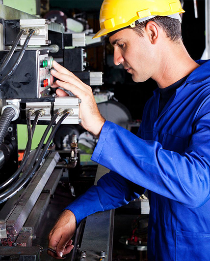Ein Mann mit einem blauen, langärmligen Pullover und einem gelben Schutzhelm steht rechts von einer technischen Anlage. Er schaut in die Anlage und drückt mit einer linken Hand einen Knopf. 