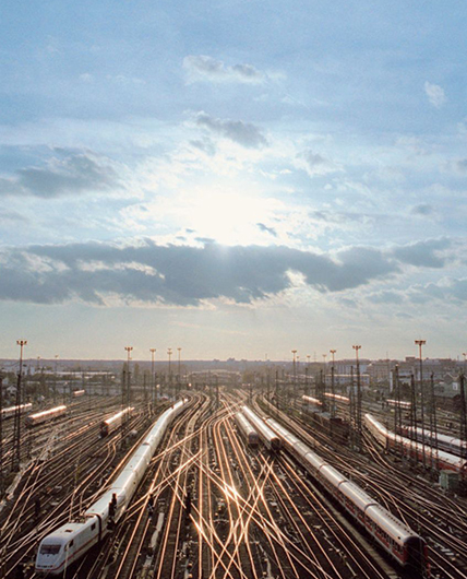 Viele parallel nebeneinander verlaufende Zuggleise. Auf einigen fahren Züge. Zwischen den Gleisen sind Strommasten. Die Gleise reichen bis zum Horizont. Im oberen Bildteil ist ein blauer Himmel mit einigen Wolken, die die Sonne verdecken. Links und rechts am Bildrand sind Häuser. 
